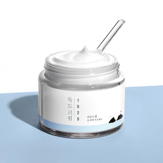 ROUND LAB's 1025 Dokdo Cream is shown with the lid off and a clear spatula in the cream, revealing its creamy texture.